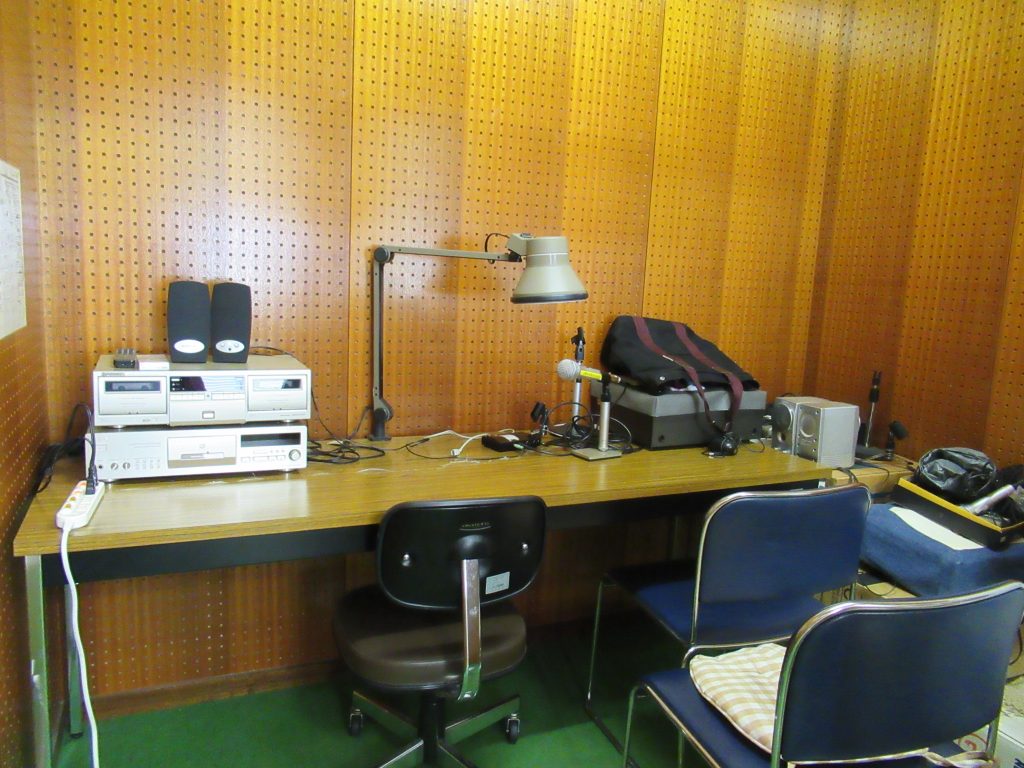 入口入った室内にある2つある録音編集スペースの中。 奥の壁付けで長机があり、机の上に各種機材があります。